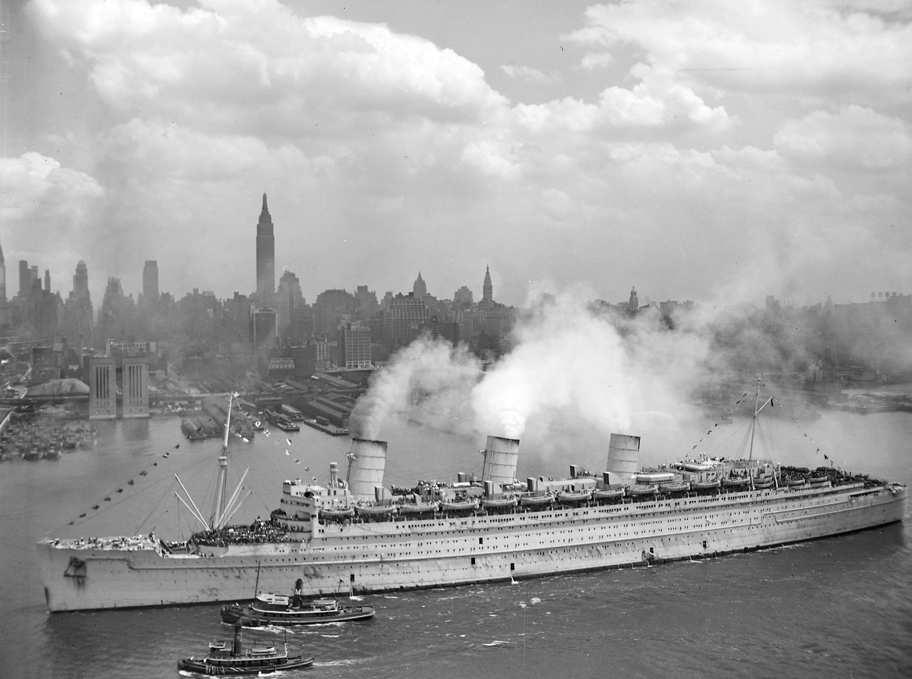Die RMS Queen Mary im Hafen von New York, 20. Juni 1945, mit vollbesetzten Decks und Schleppern